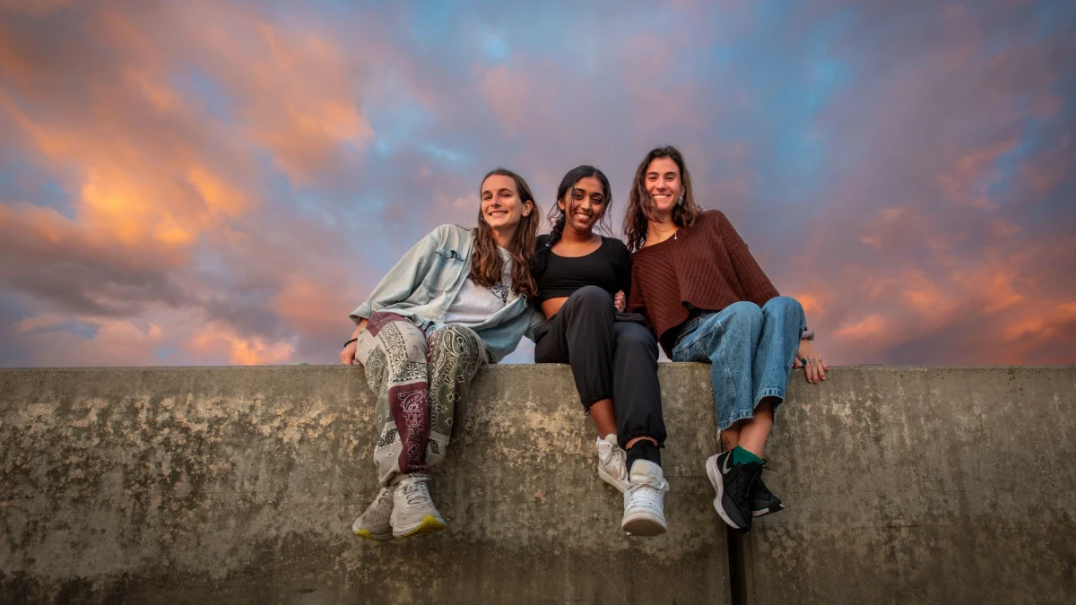 Three female students sitting on wall smiling posing for photo.